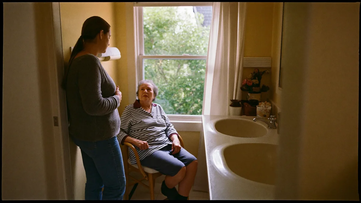 Adult daughter standing gently near a bathroom doorway, elderly parent visible seated inside on a bath chair, warm residential setting, morning light